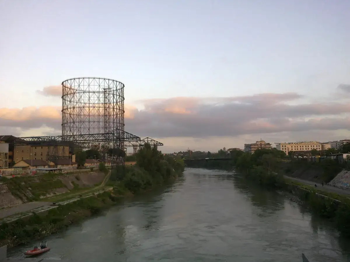 Il gasometro Roma domina il paesaggio fluviale al tramonto, con le sue strutture metalliche che si stagliano contro un cielo rosato.