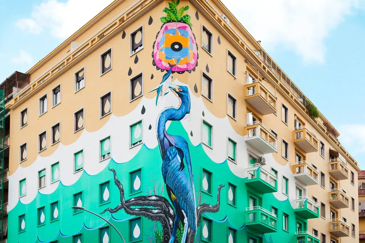 Un maestoso airone blu cattura un pesce, parte di un colorato murales ostiense che decora un edificio giallo e verde, con gocce stilizzate che cadono dal cielo.