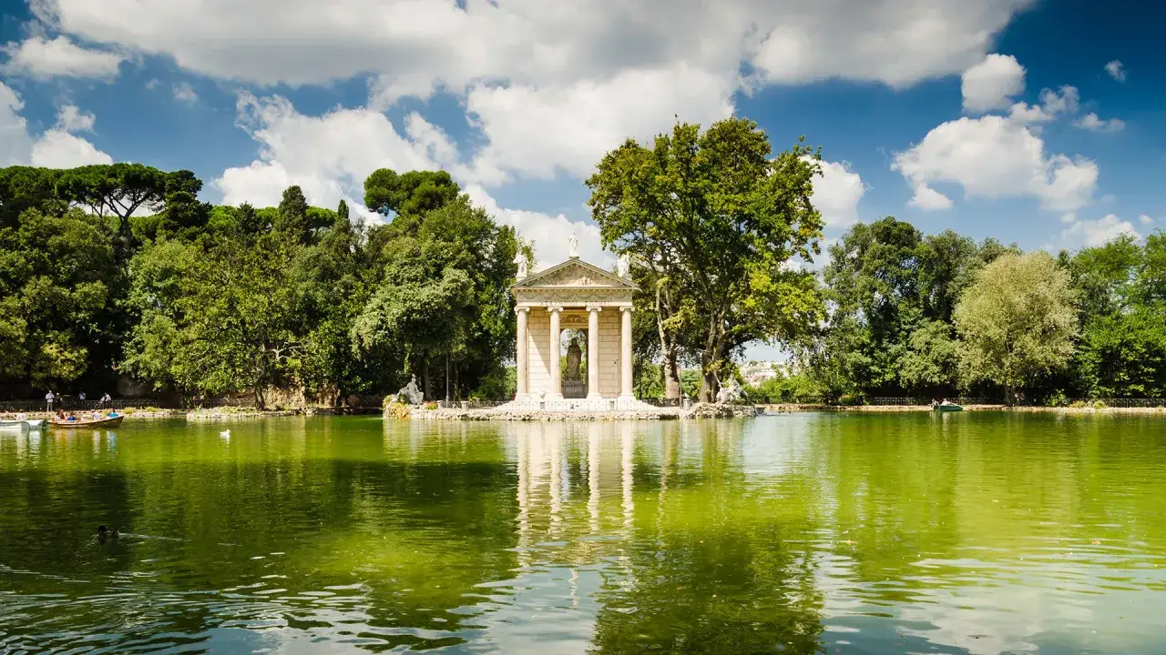 Un tempio neoclassico su un'isola nel lago dei giardini di Roma, con barche e alberi lussureggianti.