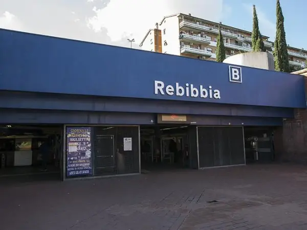 Ingresso della metro Rebibbia a Roma, con insegna blu e lettere bianche. Edifici residenziali sullo sfondo.